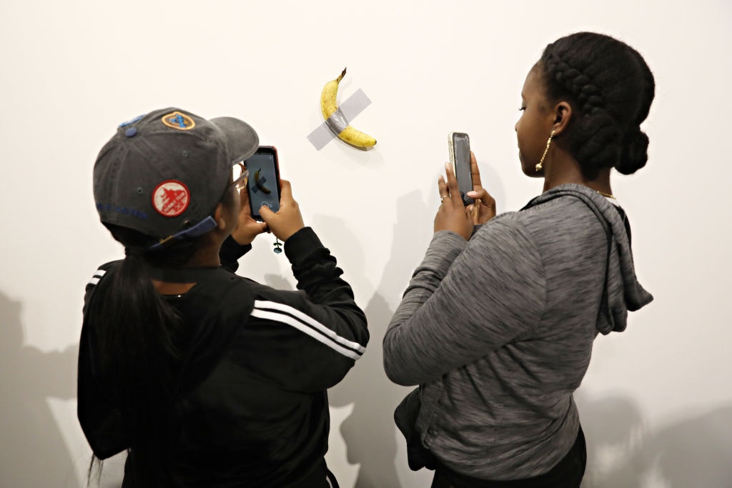 Two people take photographs with their smartphones of a banana taped to a wall.
